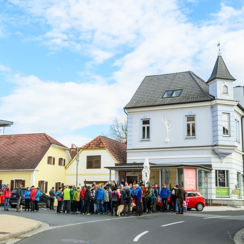 A group of people stands in front of a building with a tower. They wear jackets and backpacks. A car is parked in front of the building.
