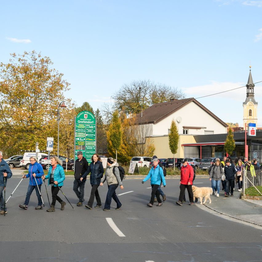 A group of people, some with walking sticks, are crossing a street in a village. Behind them are parked cars and a green signboard with 'Grafendorf'. Buildings with trees and a tower are visible in the background.