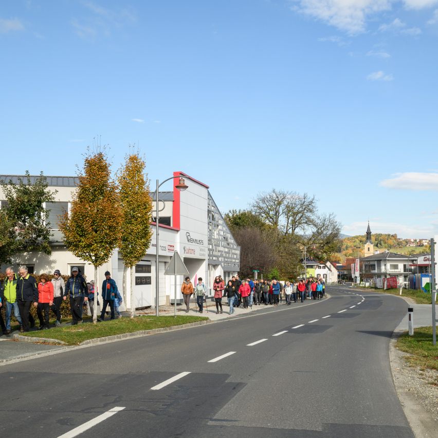 A group of people walk on the side of a road. They are dressed for cold weather. A building with a sign reading "Genuss" is nearby.