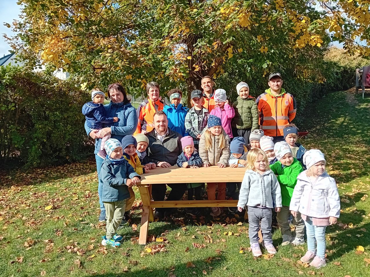 A group of adults and children, including some in winter clothing, stand behind a wooden table under a yellow-leaved tree in a park.