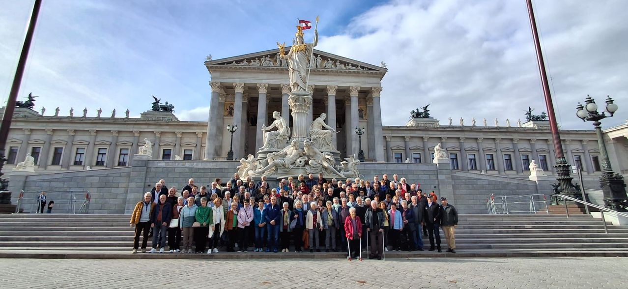 A group of elderly people stand in front of a large building with a statue and flag on top. They are wearing winter clothes and carrying bags.