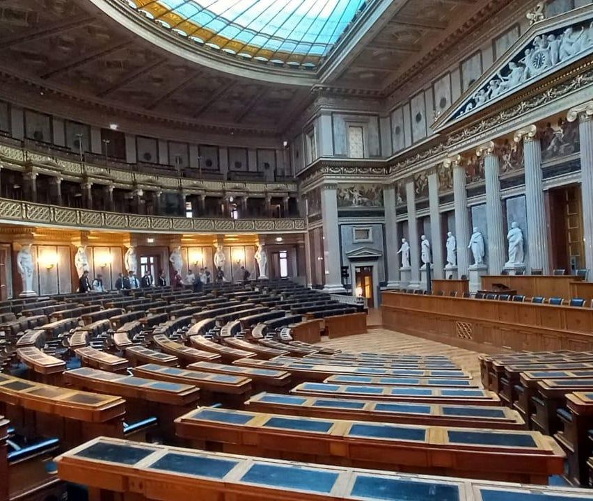 An empty parliament chamber features rows of wooden desks and seats. Above, a dome ceiling with a stained glass window. Sculptures and columns adorn the walls.