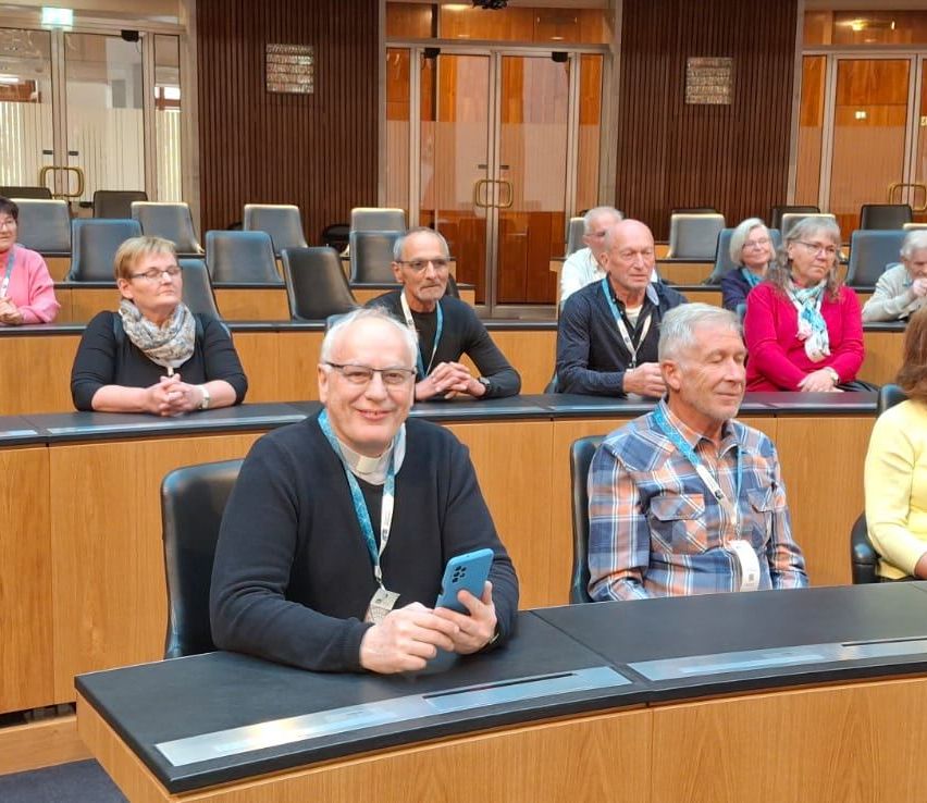 A group of older people sit in a conference room, facing the front. One person holds a cellphone.