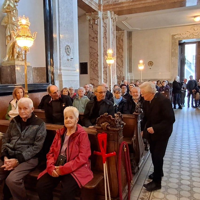 A group of elderly people sitting on pews in a church, with an older man standing in front of them. The church has decorative lamps and a statue on the wall.