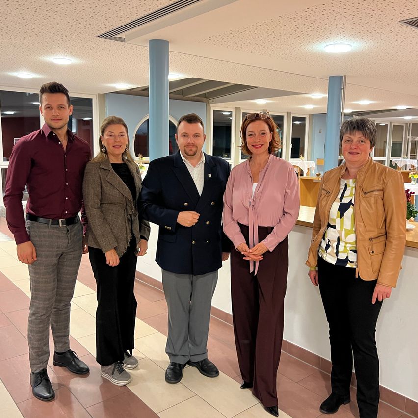 Fünf Menschen stehen in einem gut beleuchteten Raum mit gefliestem Boden und lächeln für ein Foto. Sie sind formell gekleidet. Hinter ihnen ist eine Wand mit blauen Säulen, und es gibt einen Tisch mit Blumen.