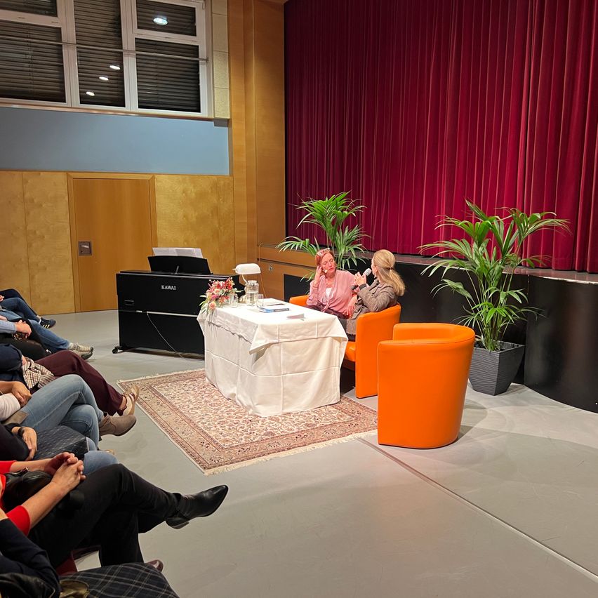 Zwei Frauen sitzen auf Stühlen und sprechen mit Mikrofonen. Ein Tisch mit einer weißen Tischdecke steht vor ihnen. Ein Klavier ist auf der linken Seite. Es gibt Topfpflanzen auf der Bühne. Mehrere Menschen sitzen auf dem Boden.