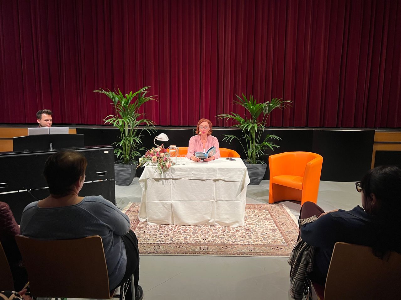 Eine Frau in einem rosa Shirt sitzt an einem Tisch und liest ein Buch. Hinter ihr ist ein roter Vorhang und ein Teppich. Zwei Personen sitzen in Stühlen und lauschen.