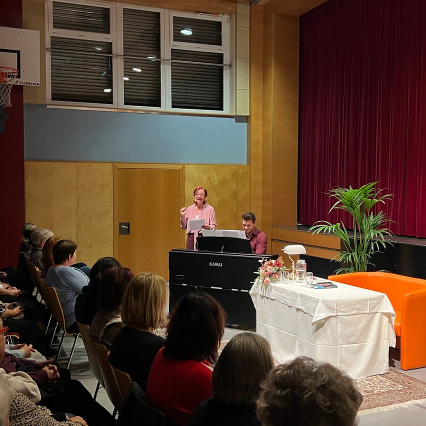 Eine Frau spricht vor einem Klavier, während ein Mann in einem Raum mit rotem Vorhang und Basketballkorb spielt. Mehrere Personen sitzen auf Stühlen.