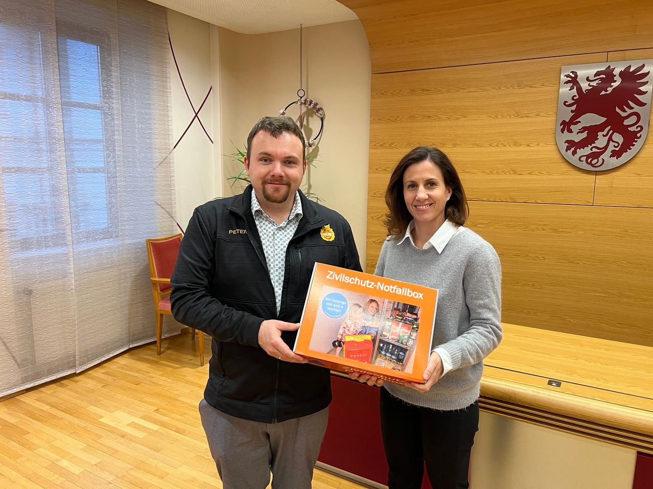 Ein Mann und eine Frau stehen in einem Raum, lächeln und halten eine Schachtel mit der Aufschrift 'Zivischutz-Notfallbox'. Der Mann trägt eine Jacke und die Frau einen grauen Pullover. Hinter ihnen befindet sich eine Holzwand und ein Stuhl. Der Raum hat Holzboden.