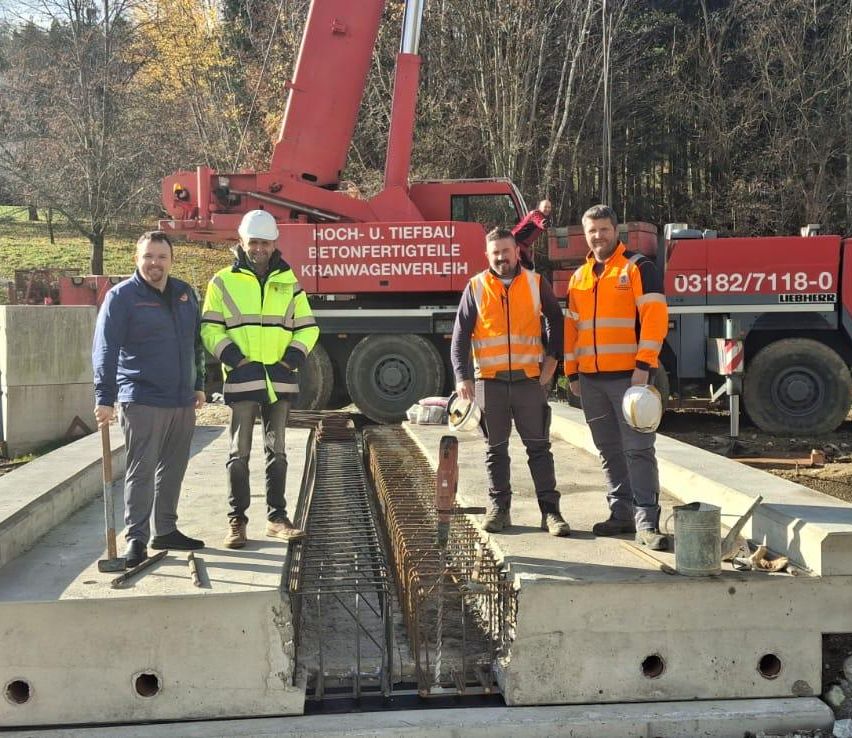 Vier Männer in Warnwesten posieren vor einer Betonstruktur auf einer Baustelle, mit einem roten Kran und einem Fahrzeug im Hintergrund.