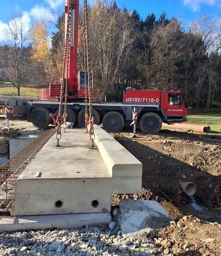 Ein großer roter Kran hebt eine Betonplatte an einer Baustelle. Der Kran hat die Nummer 03182/7118-0. In der Nähe gibt es einen Haufen Kies und eine Wasserleitung.