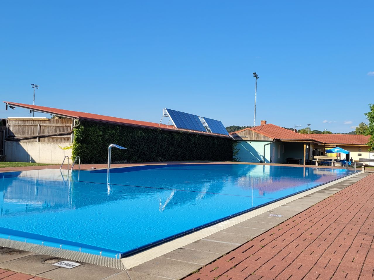 Ein Freischwimmbecken mit blauer Wasseroberfläche und einem gepflasterten Gehweg. Solarmodule sind auf dem Dach montiert.