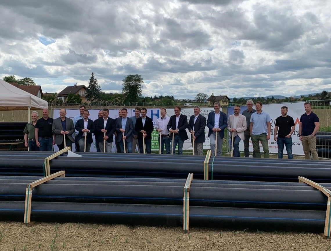 Gruppe von Männern posiert hinter Rohren auf einer Baustelle. Einige halten Schaufeln. Dahinter ist ein Banner sichtbar.