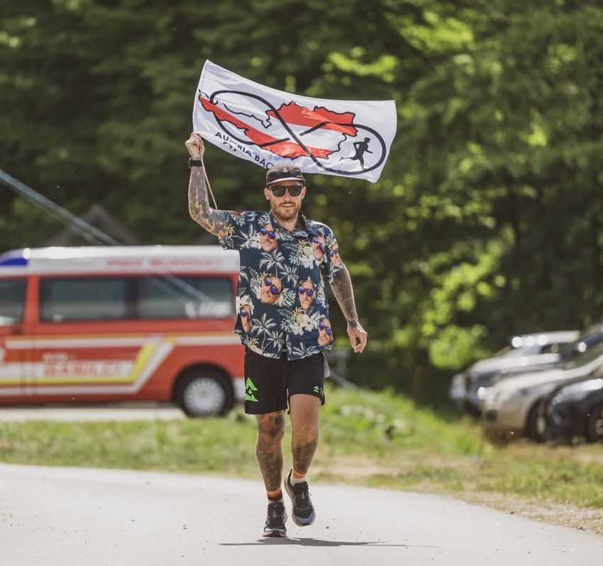 A man runs a marathon in the forest, holding a flag with an Austrian map. A red van and parked cars are in the background.