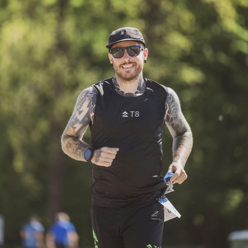 A man with tattoos runs in the forest, wearing a black shirt and hat. He holds a bottle and a paper in his hand.