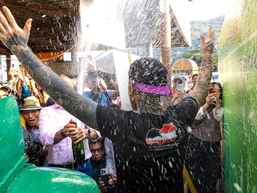 A man in a black shirt celebrates in a crowd, spraying champagne, with a green object behind him and people taking photos.