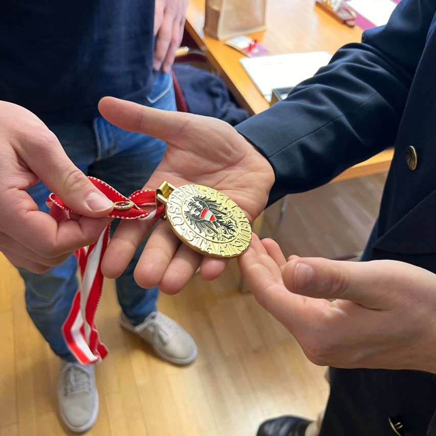 Two people hold a golden medal with a red ribbon, possibly in a classroom. A desk with a bag and papers is in the background.
