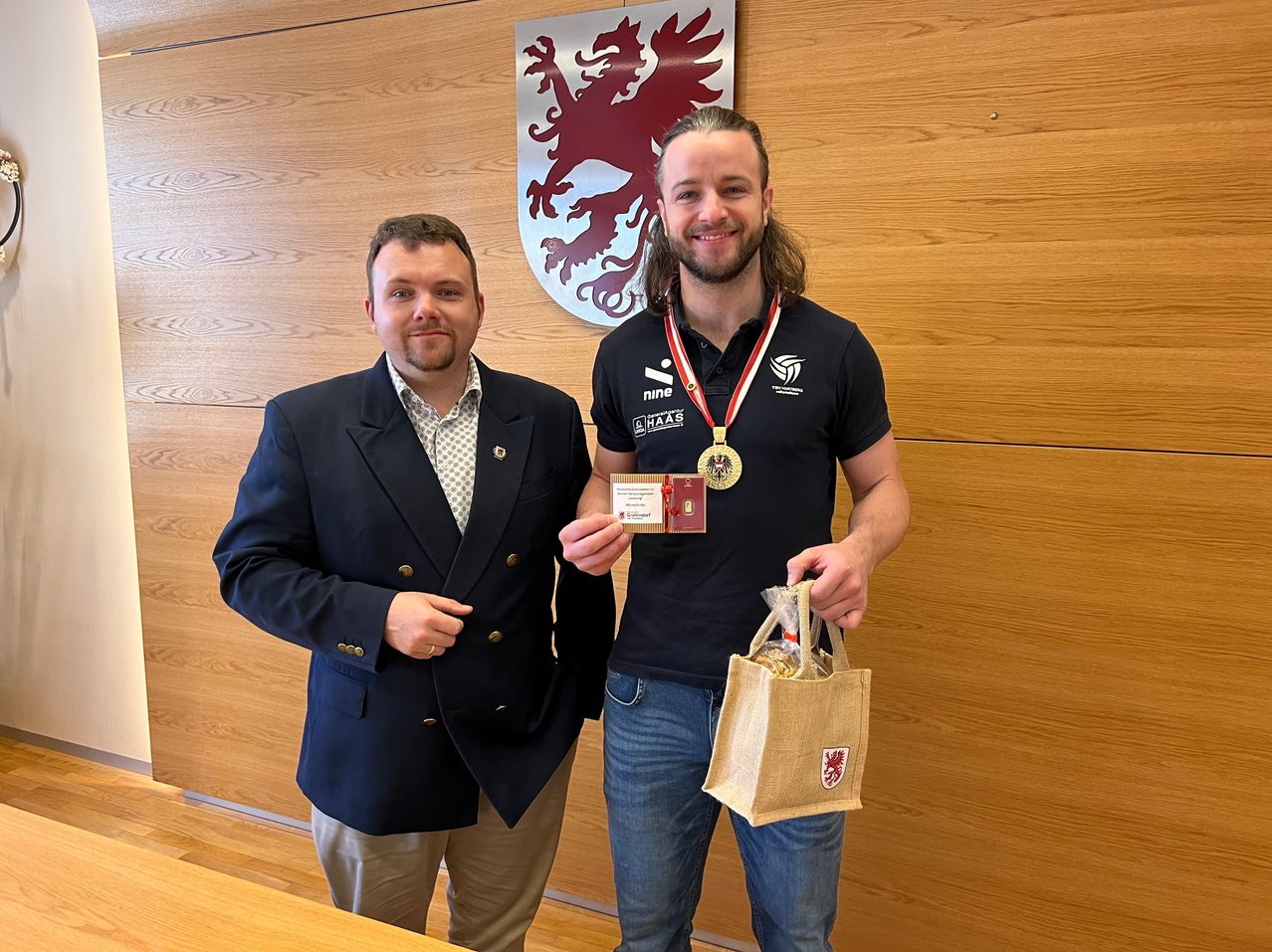 Two men standing, one in a suit holding a card, the other in a jersey with a medal and a bag. Behind them is a wall with a red lion emblem.