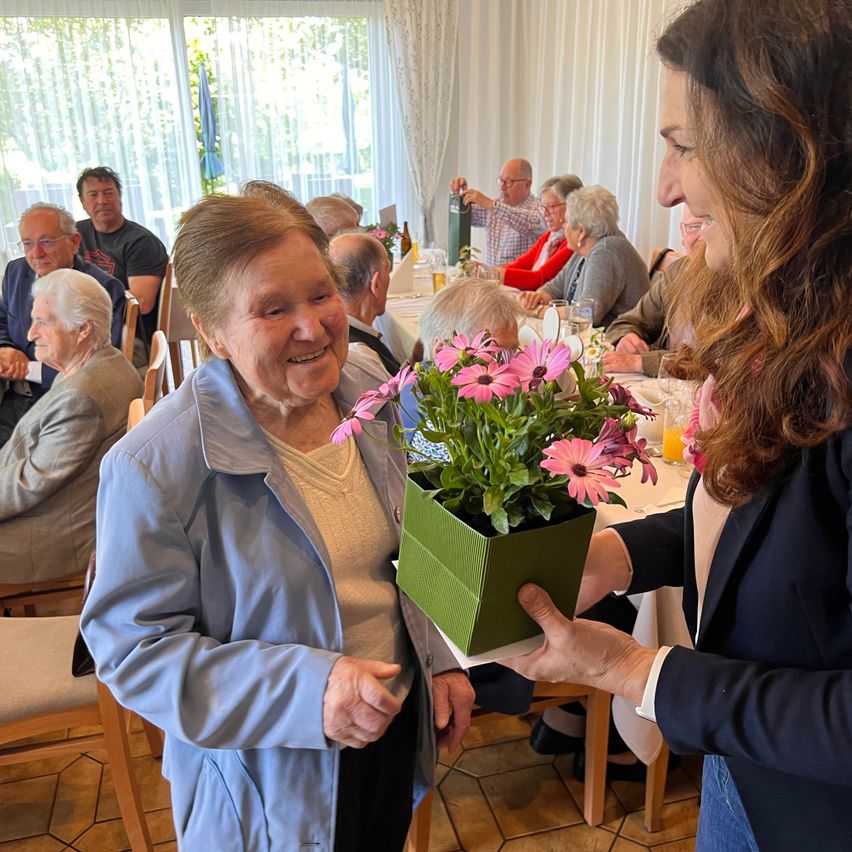 An elderly woman with short hair smiles while holding a green potted flower arrangement, another woman hands her the flowers. Other people are sitting and talking in the background.