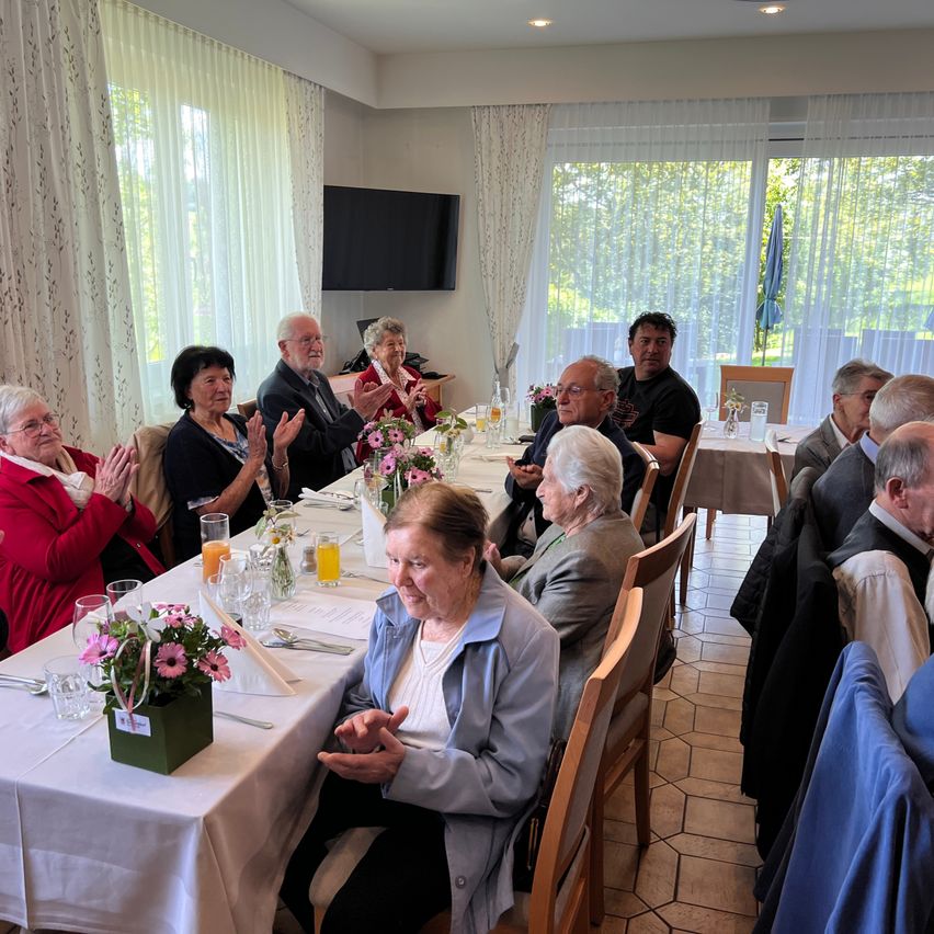 A group of older people sit at a long table with flowers, clapping. The room has white walls, a large window, and a TV on the wall.