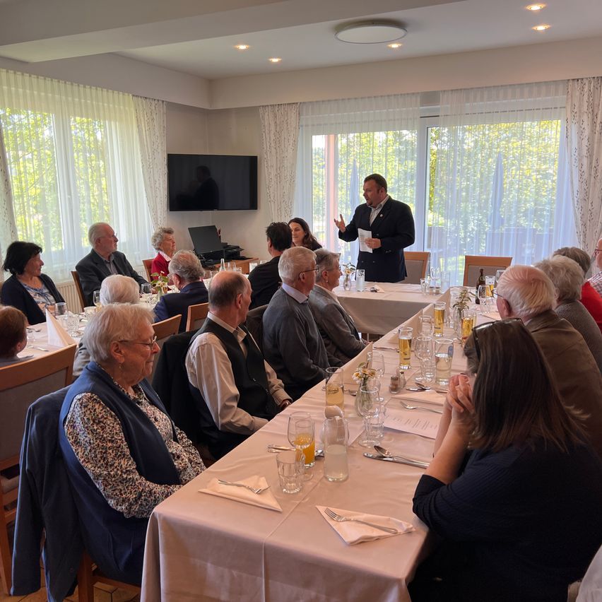 A group of elderly people sit at tables in a dining room, listening to a man standing at the front giving a speech. There are wine glasses, a television, and large windows in the room.