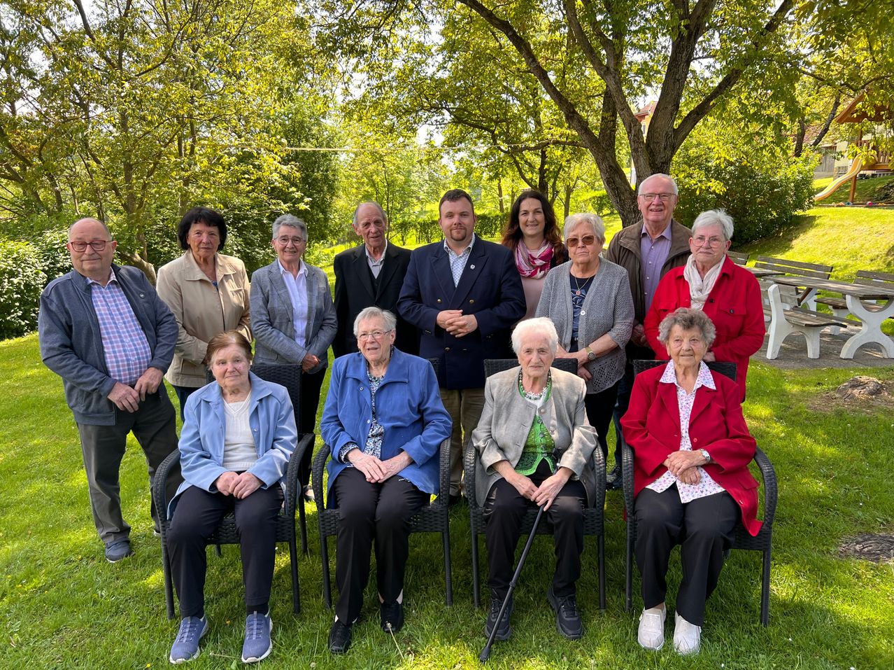 A group of elderly people are posing for a photo in a park. Some are seated in chairs, while others are standing. There are trees in the background.
