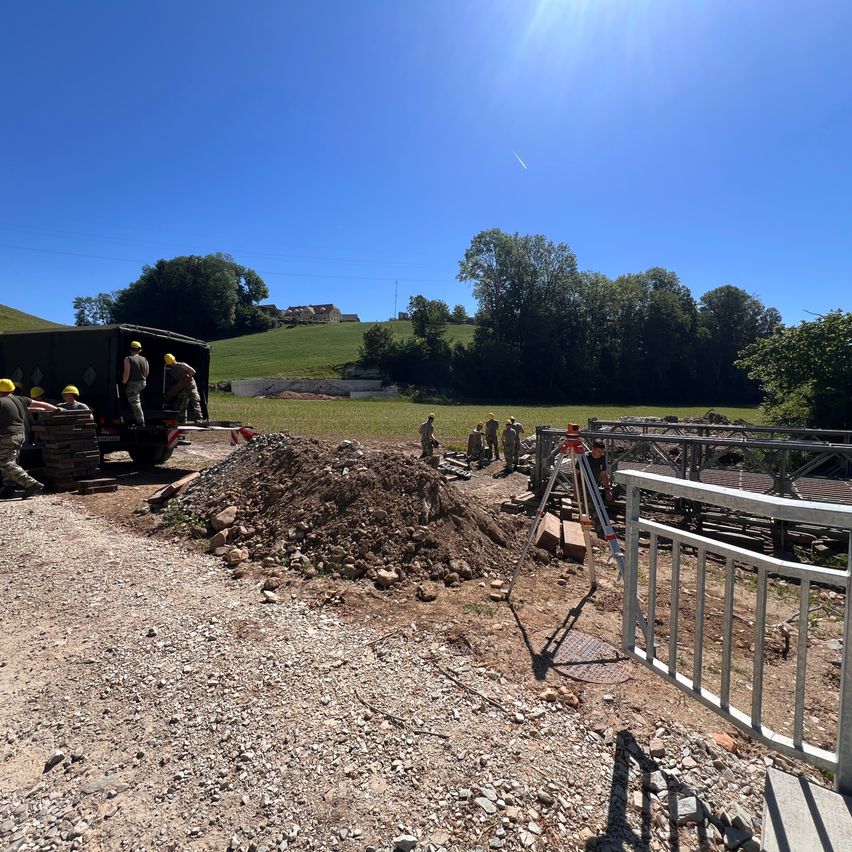 Workers in hard hats unload materials from a truck. A pile of dirt and rocks is nearby. In the background, there is a fence and a hillside with greenery.