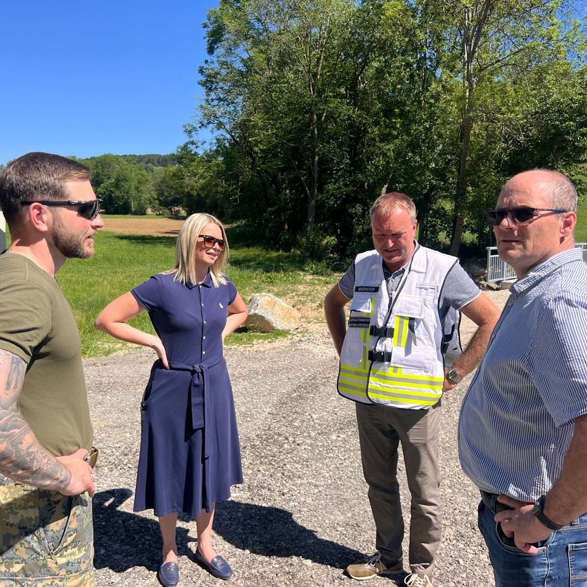 Four people stand in an outdoor setting, facing each other. A woman in a navy dress stands between two men in casual wear, while another man wears a reflective vest. The background features greenery and a clear blue sky.