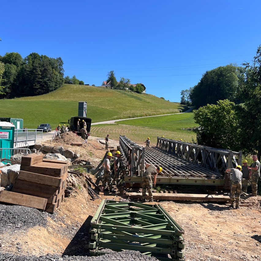 A group of workers in military uniforms are constructing a bridge over a dirt path. Piles of wooden boards and metal structures are on the ground. In the background, a car and a house are visible.