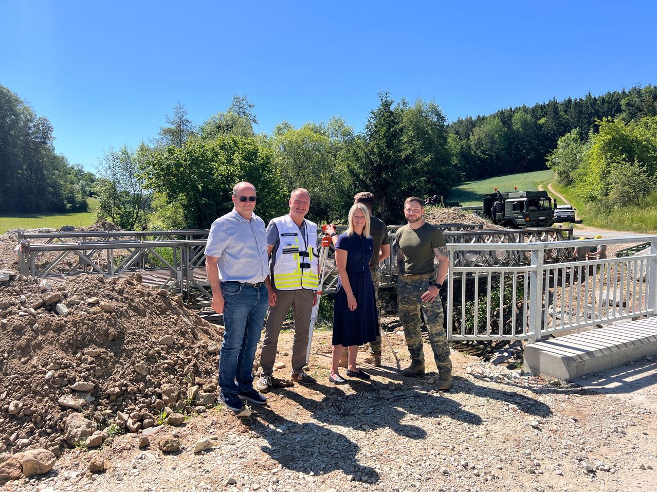 Five people stand in front of a white fence in a construction area. Behind them are piles of gravel and a bridge. In the distance, a truck is parked on the road.