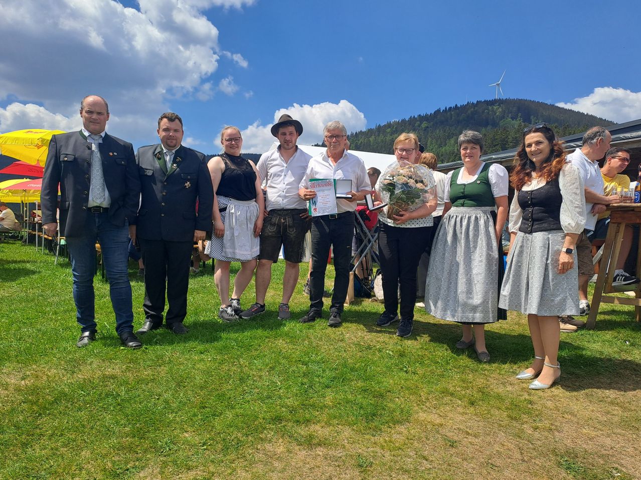 A group of people are standing on a grassy field, possibly at a festival. A woman holds flowers and a certificate. A windmill is in the background.