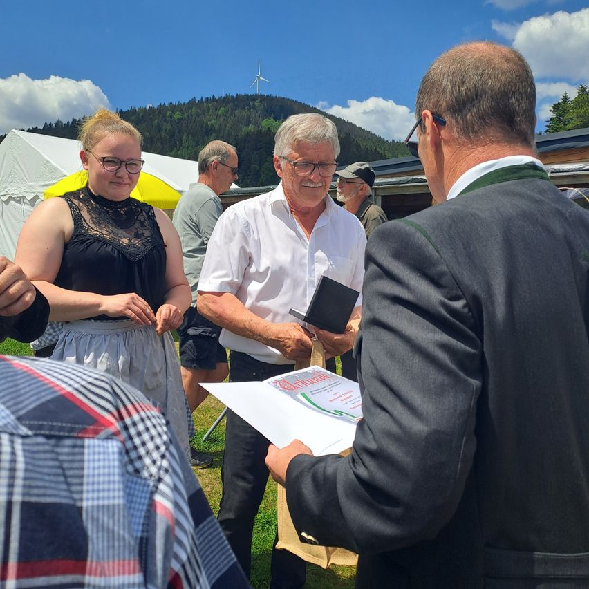 A group of people stands outside with mountains in the background. A man in glasses holds a folder and looks at a document, while another man looks on. A woman in a black dress and glasses smiles.