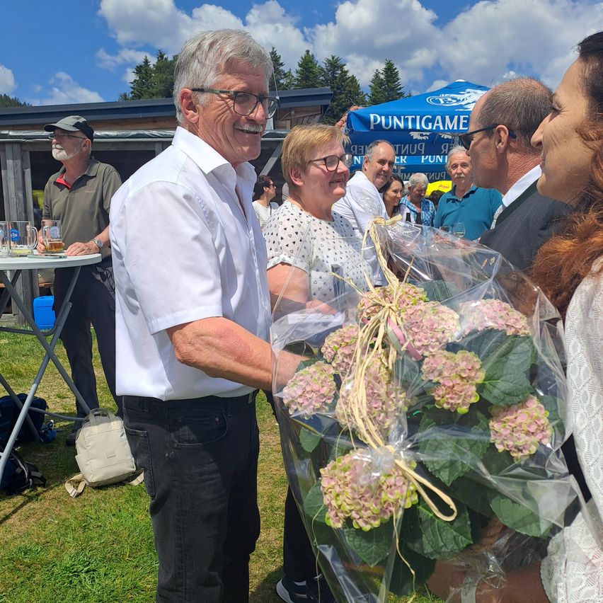 An older man with glasses holds a bouquet of pink flowers, smiling and standing outside. Behind him, a woman with glasses smiles, and several people stand under a tent.