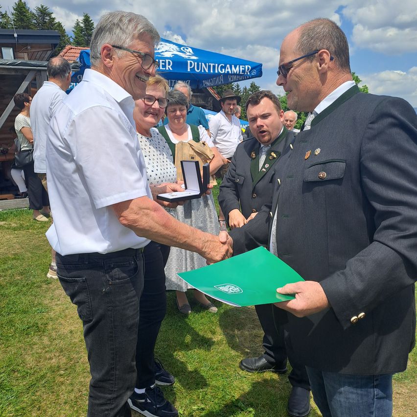 A man in formal attire shakes hands with another man holding a green folder. A woman smiles beside them. They are surrounded by people and a blue tent.