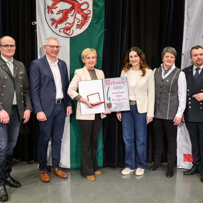 Eine Gruppe von Menschen steht vor einer grünen Flagge mit einem roten Löwenwappen. Eine Frau hält ein Zertifikat.