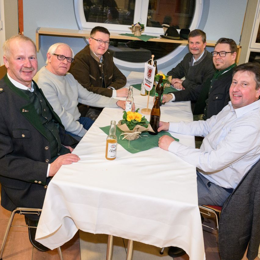 Sechs Männer sitzen um einen Tisch herum. Sie alle lächeln und scheinen eine gute Zeit zu haben. Sie tragen Mäntel, und auf dem Tisch stehen Flaschen und eine Blumendekoration. Der Raum hat ein großes kreisförmiges Fenster.