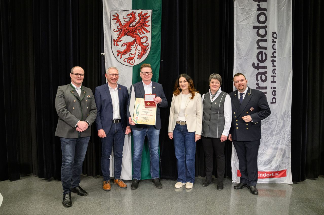 Sechs Menschen stehen vor einem Hintergrund mit einer Flagge. Ein Mann hält ein gerahmtes Zertifikat.
