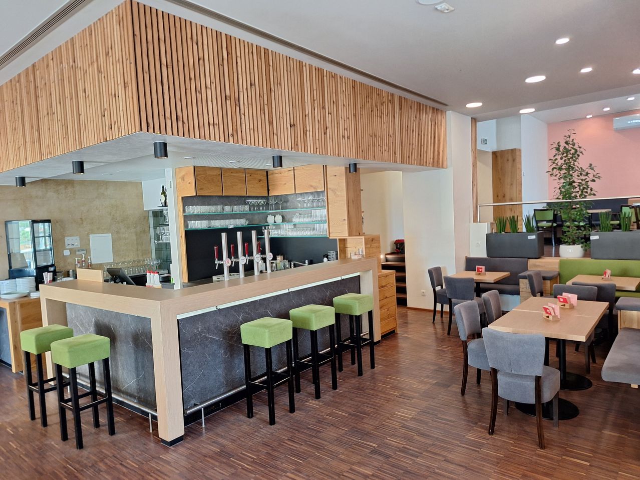 An empty restaurant with wooden floors, a bar counter with stools, and tables with chairs. The bar counter has a marble top and wooden cabinets.