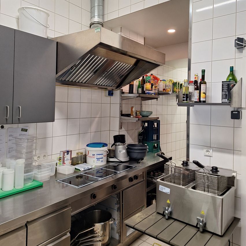 A commercial kitchen with stainless steel appliances, a large stove, and a deep fryer. Bottles and containers are on shelves. A white bucket is on a cabinet.