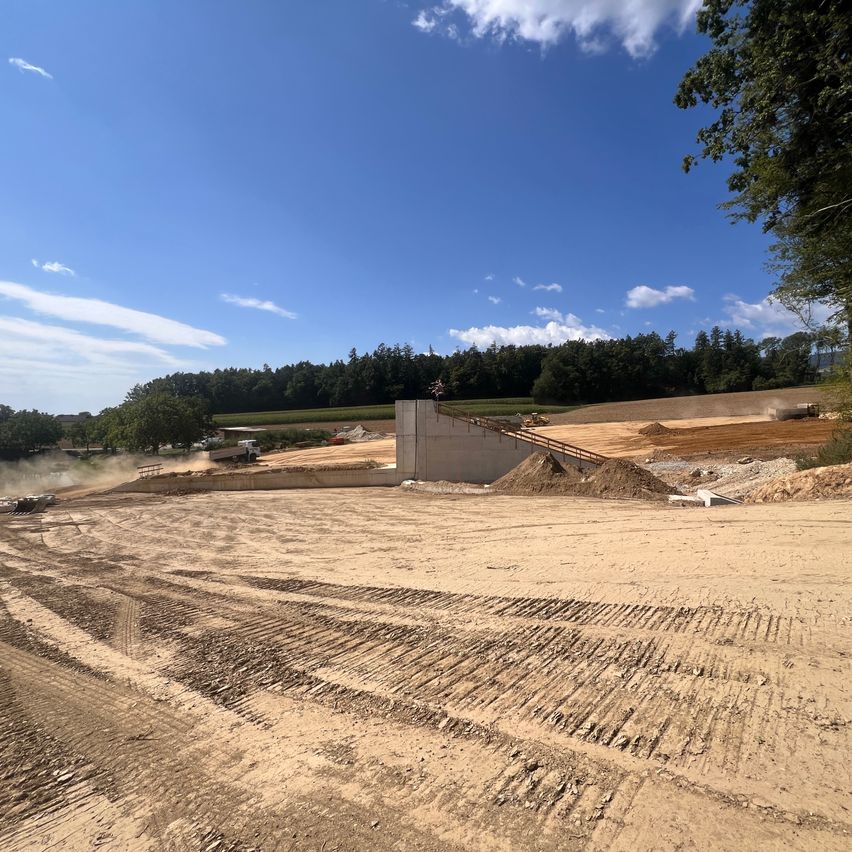 Eine Baustelle mit einer Betonmauer, Erde und Baumaschinen unter einem klaren blauen Himmel mit einigen Wolken.