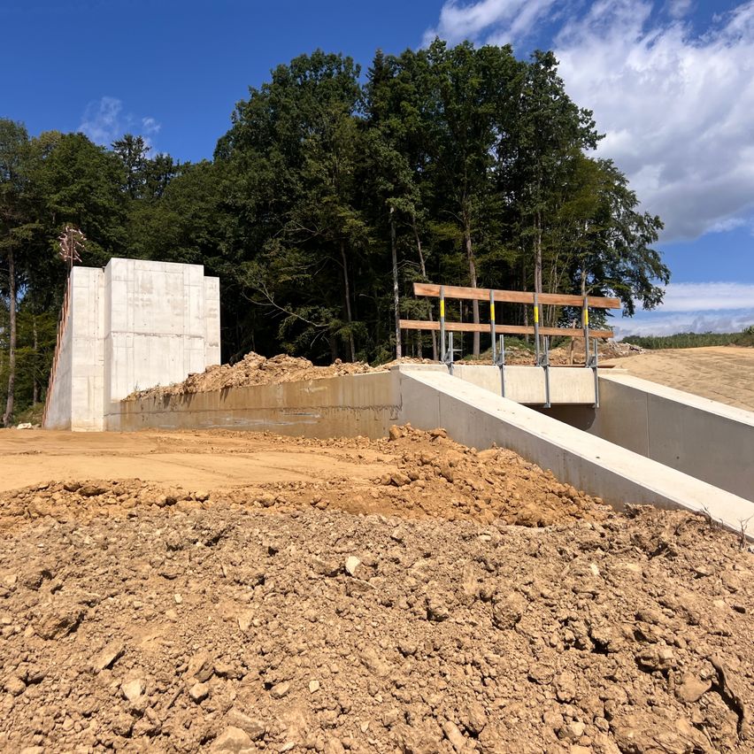 Eine Betonstruktur im Bau mit einer hölzernen Stütze in einem Außenbereich mit Erde und Bäumen im Hintergrund.
