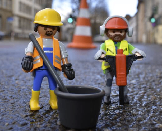 Zwei Playmobil-Bauarbeiter stehen auf einer nassen Straße, einer hält ein Werkzeug und der andere eine Bohrmaschine. Vor ihnen steht ein schwarzer Eimer. Dahinter befindet sich ein Verkehrskegel und ein Gebäude.