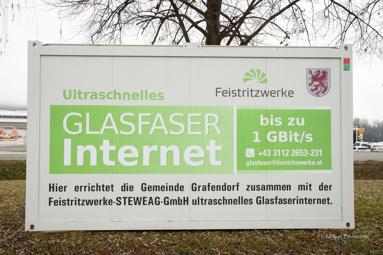 Ein weißes Schild mit grünem Text bewirbt ultraschnelle Internetdienste von Glasfaser Internet. Es enthält ein Logo, Kontaktdaten und ein Wappen.
