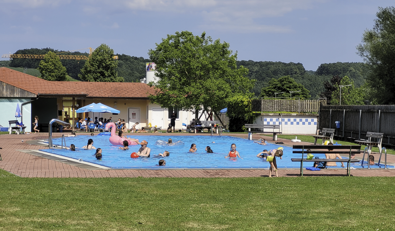Eine Gruppe von Kindern und Erwachsenen schwimmt in einem großen Freibad mit einem blauen Sonnenschirm und einem Baum im Hintergrund.