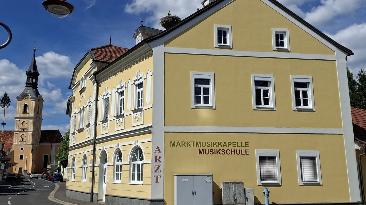 Ein gelbes Gebäude mit weißen Begrenzungen hat mehrere Fenster und ein Dach mit einem Vogelnest. Es steht 'Marktmusikkapelle Musikschule' an der Seite.