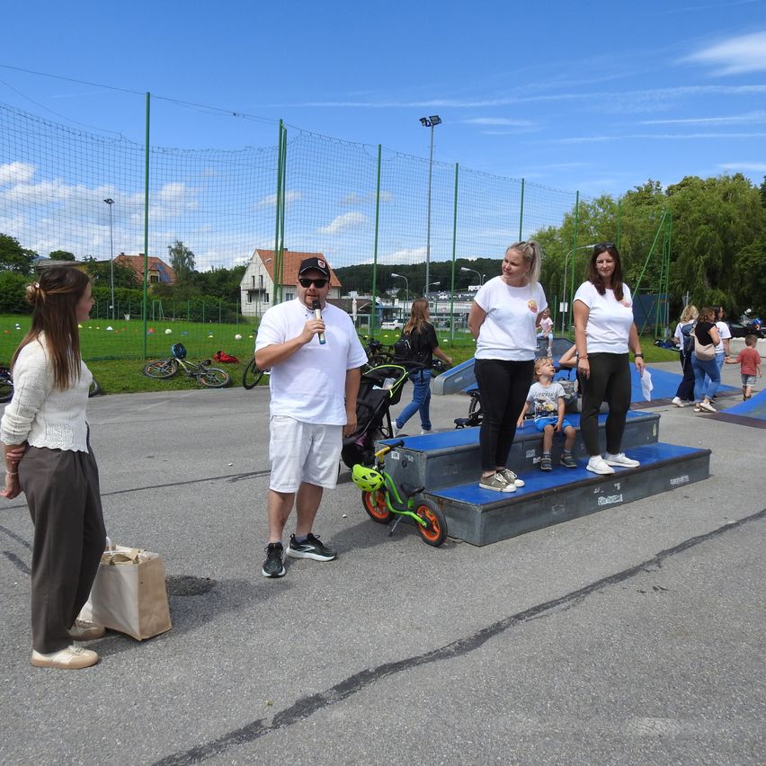 Eine Gruppe von Menschen steht auf einer Plattform mit einem Kind darauf sitzend. Ein Mann hält ein Mikrofon und andere schauen umher. Fahrräder und ein Kinderwagen sind in der Nähe geparkt. In der Ferne stehen Häuser und ein Netzzaun.