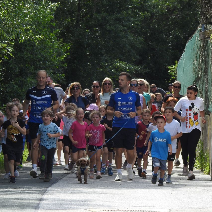 Eine Gruppe von Menschen und Kindern geht mit einem Hund an der Leine auf einer Straße. Bäume und ein Zaun sind im Hintergrund zu sehen.