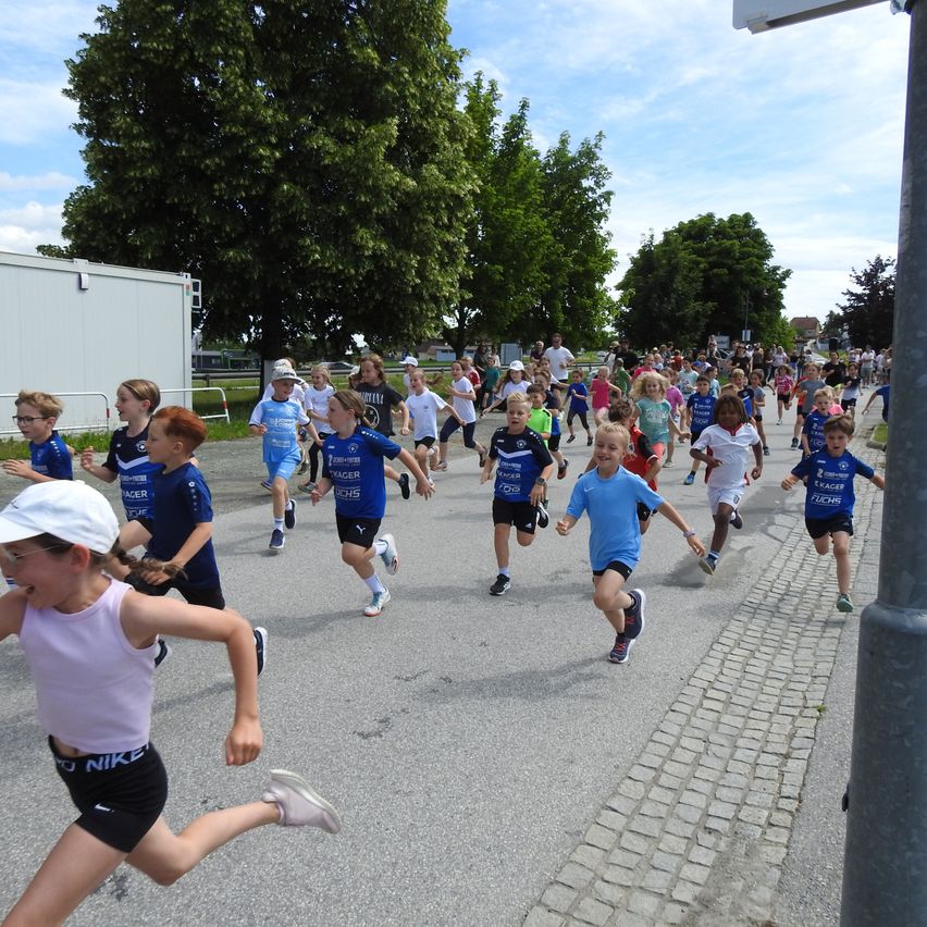 Eine große Gruppe von Kindern läuft in einem Rennen, trägt Uniformen mit Text und Zahlen. Einige tragen Brillen, Mützen und Turnschuhe. Das Rennen findet auf einer asphaltierten Straße statt, mit Bäumen und Gebäuden im Hintergrund.