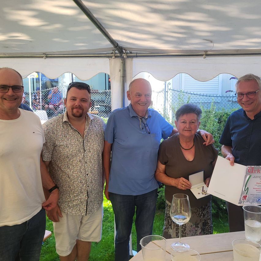 Fünf Personen posieren für ein Foto in einem Zelt. Eine Frau hält eine Urkunde, und ein Mann ein Glas. Sie alle lächeln, und auf dem Tisch stehen Gläser und Becher.