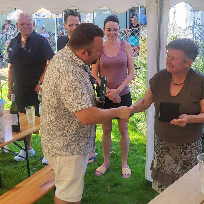 Ein Mann übergibt eine Auszeichnung an eine Frau im Garten, mit mehreren Menschen hinter ihnen. Die Frau hält ein kleines Buch.